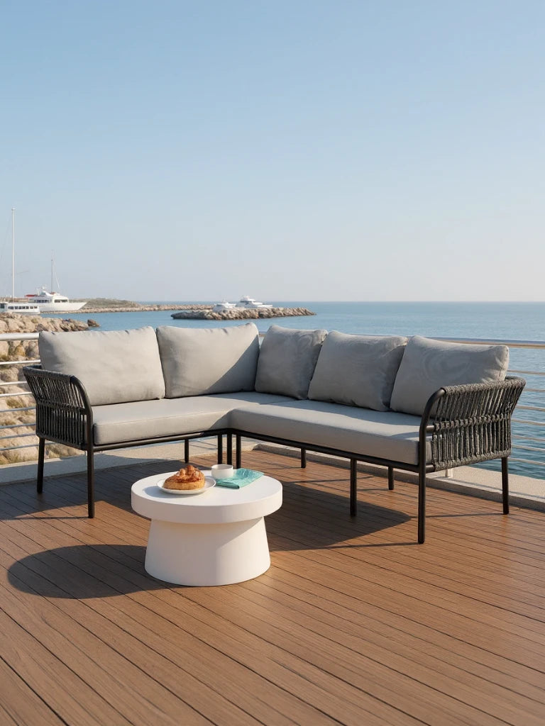 Malaga Garten Ecklounge Set (4-Sitzer) auf einer Holzterrasse mit Blick auf Wasser. Enthält ein modernes L-förmiges Sofa mit hellgrauen Kissen und einem weißen, runden Couchtisch mit Gebäck und Tasse.