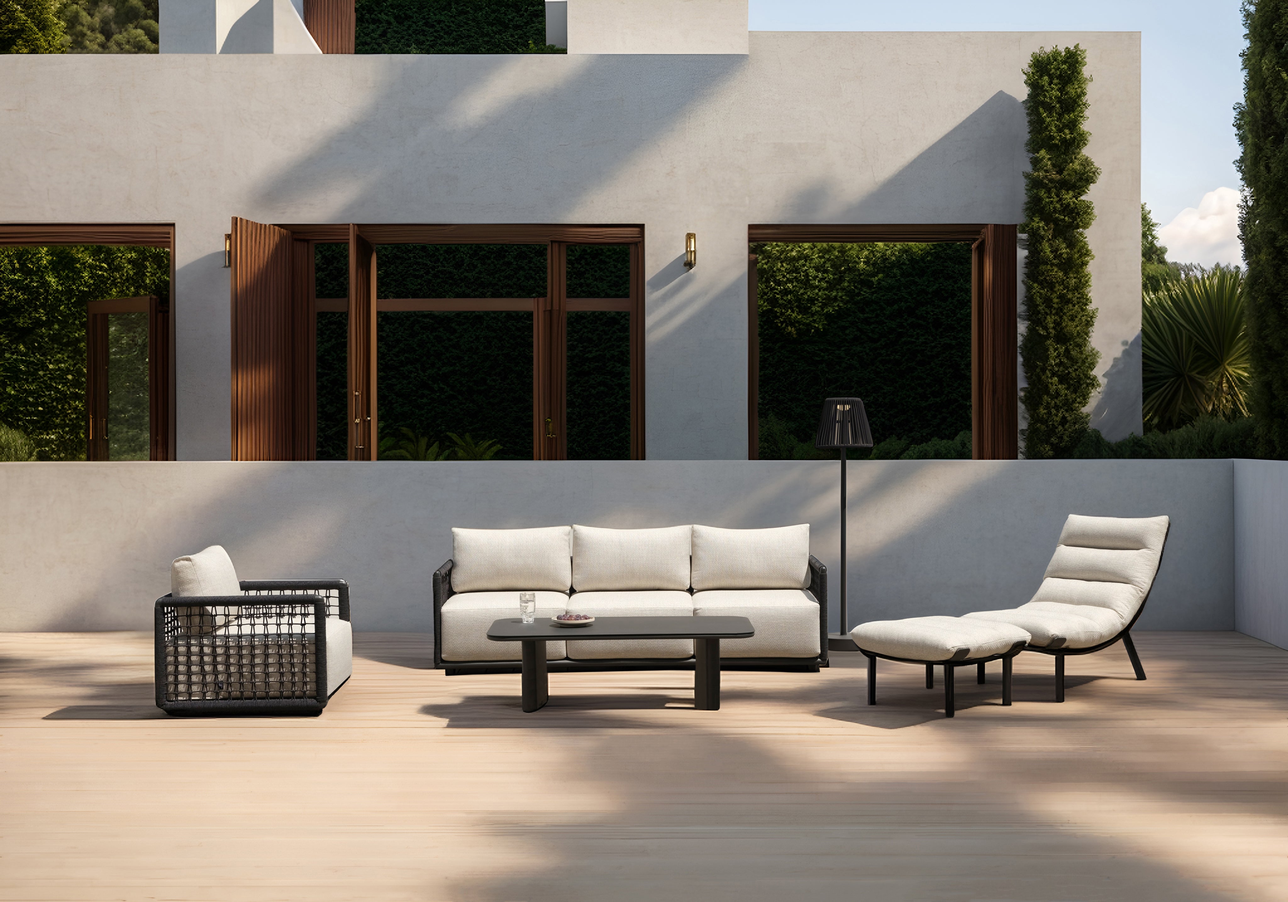 Mykonos Garten Ecklounge Set auf einer Holzterrasse: schwarzer Rahmen, cremeweiße Polster, bestehend aus einem Sessel, Dreisitzer-Sofa, Couchtisch mit Glas, Chaiselongue und Stehlampe vor grünen Hecken.