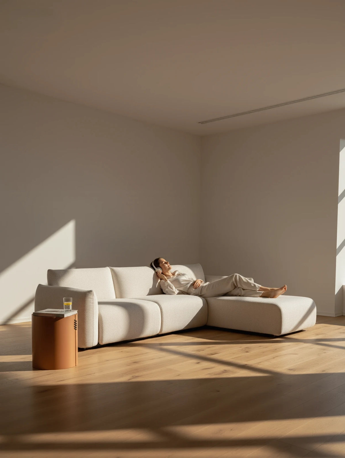 Frau entspannt sich auf dem BabyMax Einzelartikel Liege mit einer Armlehne, einem modularen, cremefarbenen Sofa, während sie Kopfhörer trägt; neben ihr ein Tisch mit Getränk und Magazin.