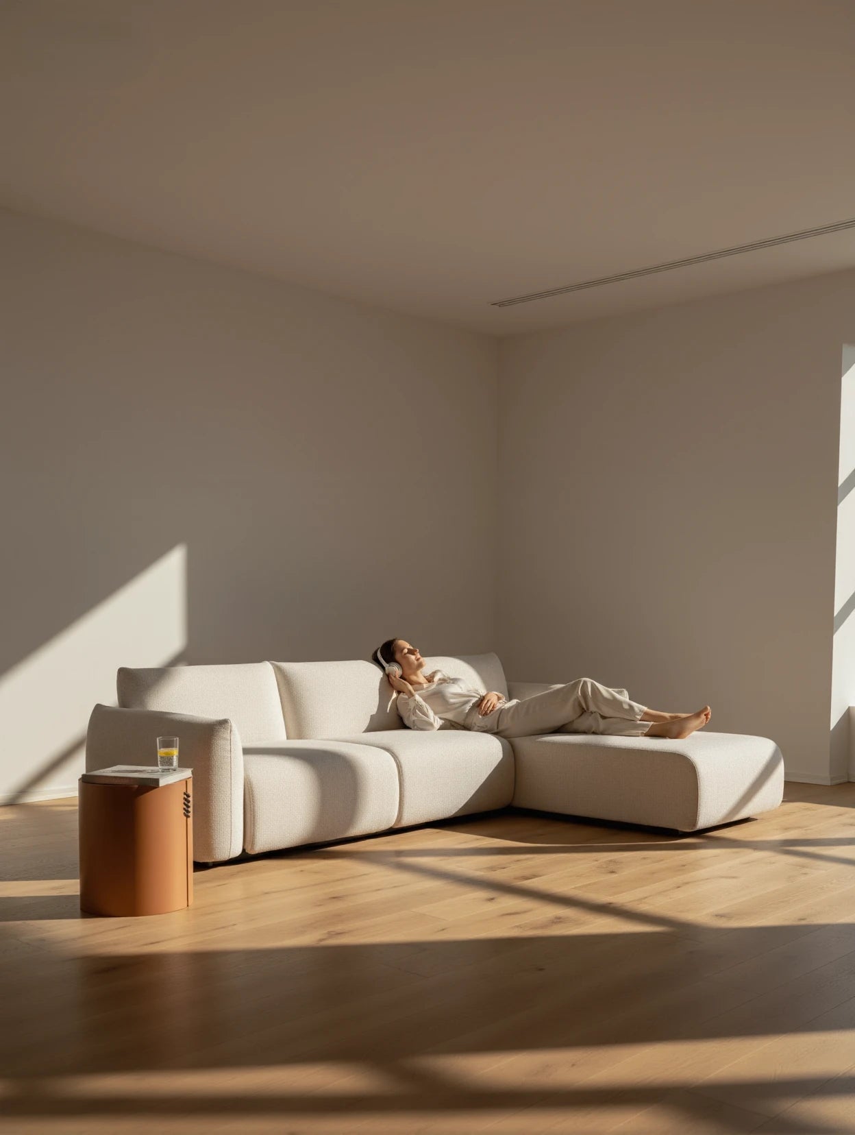 Babymax L-Lounge Set, 2er-Set: Ein modularer, cremefarbener L-förmiger Sofa in einem minimalistischen Raum. Eine Person entspannt darauf mit Kopfhörern, betont durch sanfte Sonneneinstrahlung und geometrische Schatten.