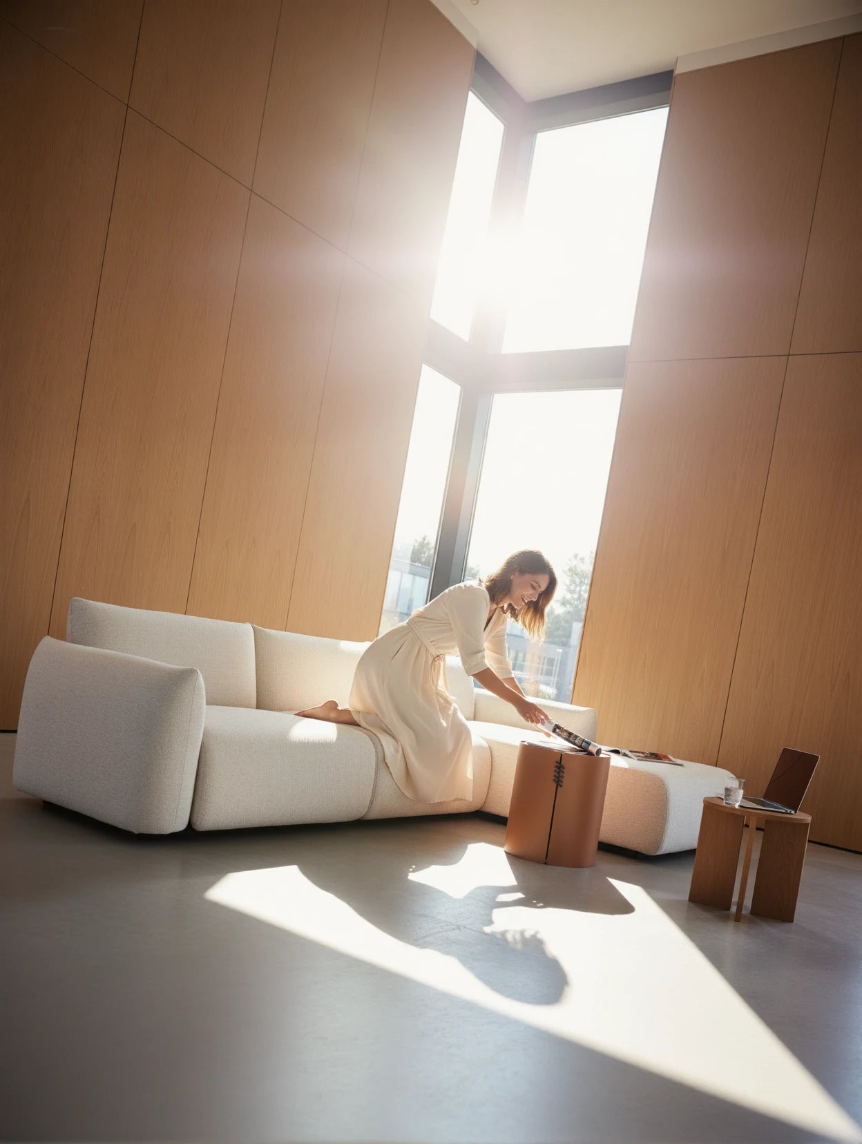 Babymax L-Lounge Set, 2er-Set im minimalistischen Wohnzimmer, platziert vor warmen Holzpaneelen, mit einer Frau auf dem Sofa, die sich zu einem kleinen Tisch mit Laptop und Wasser neigt.