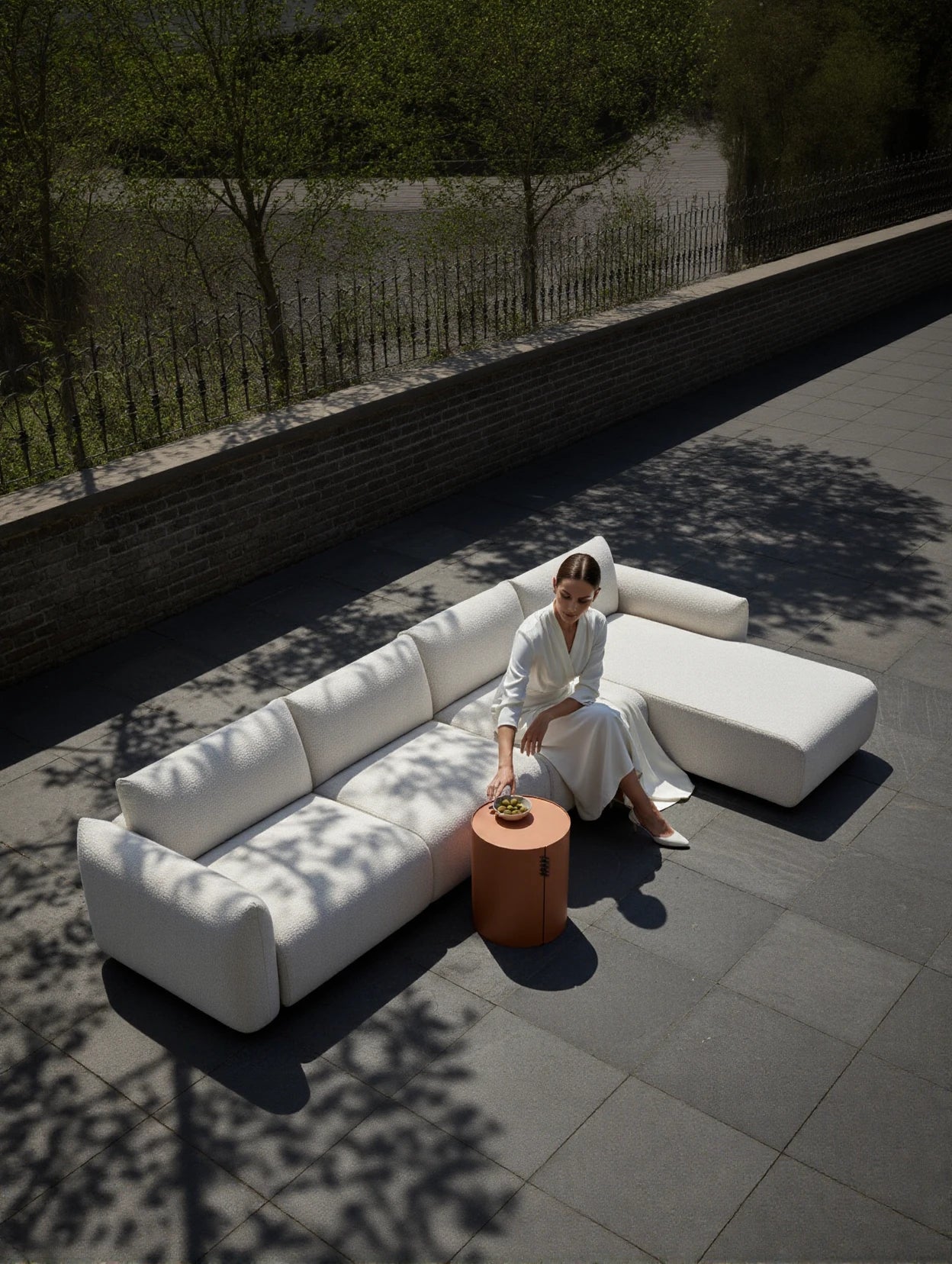 Babymax L-Lounge Set, 3er-Set: Minimalistisches, weiß gepolstertes Outdoor-Sofa mit Rechts-Chaise, auf einer großen Terrasse platziert. Eine Frau greift nach einem Terrakotta-Beistelltisch mit einer Schale grüner Früchte.
