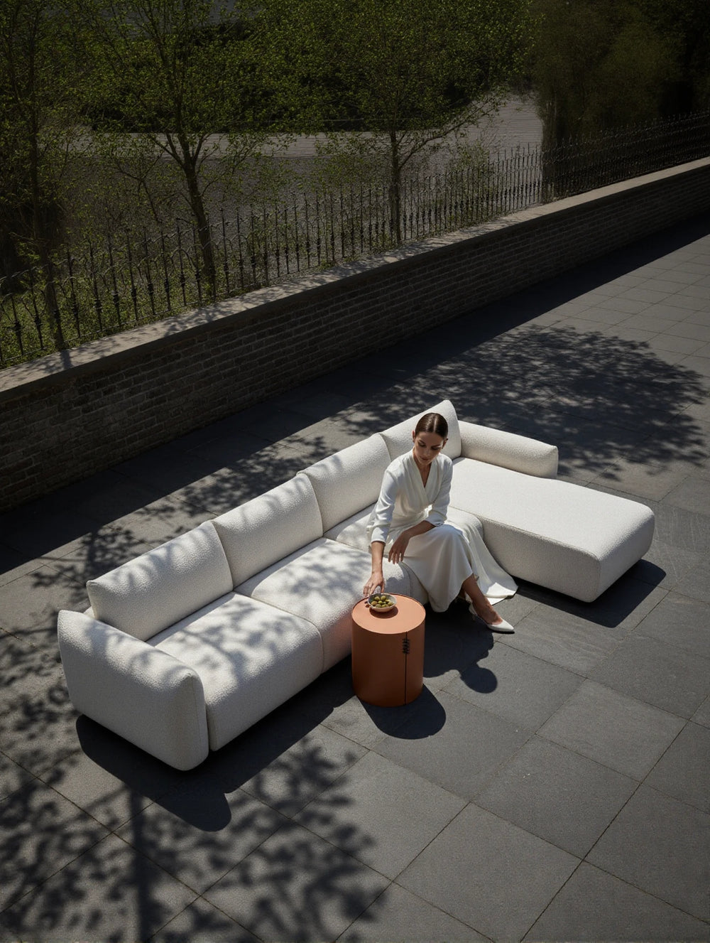 Babymax L-Lounge Set, 3er-Set: Minimalistisches, weiß gepolstertes Outdoor-Sofa mit Rechts-Chaise, auf einer großen Terrasse platziert. Eine Frau greift nach einem Terrakotta-Beistelltisch mit einer Schale grüner Früchte.