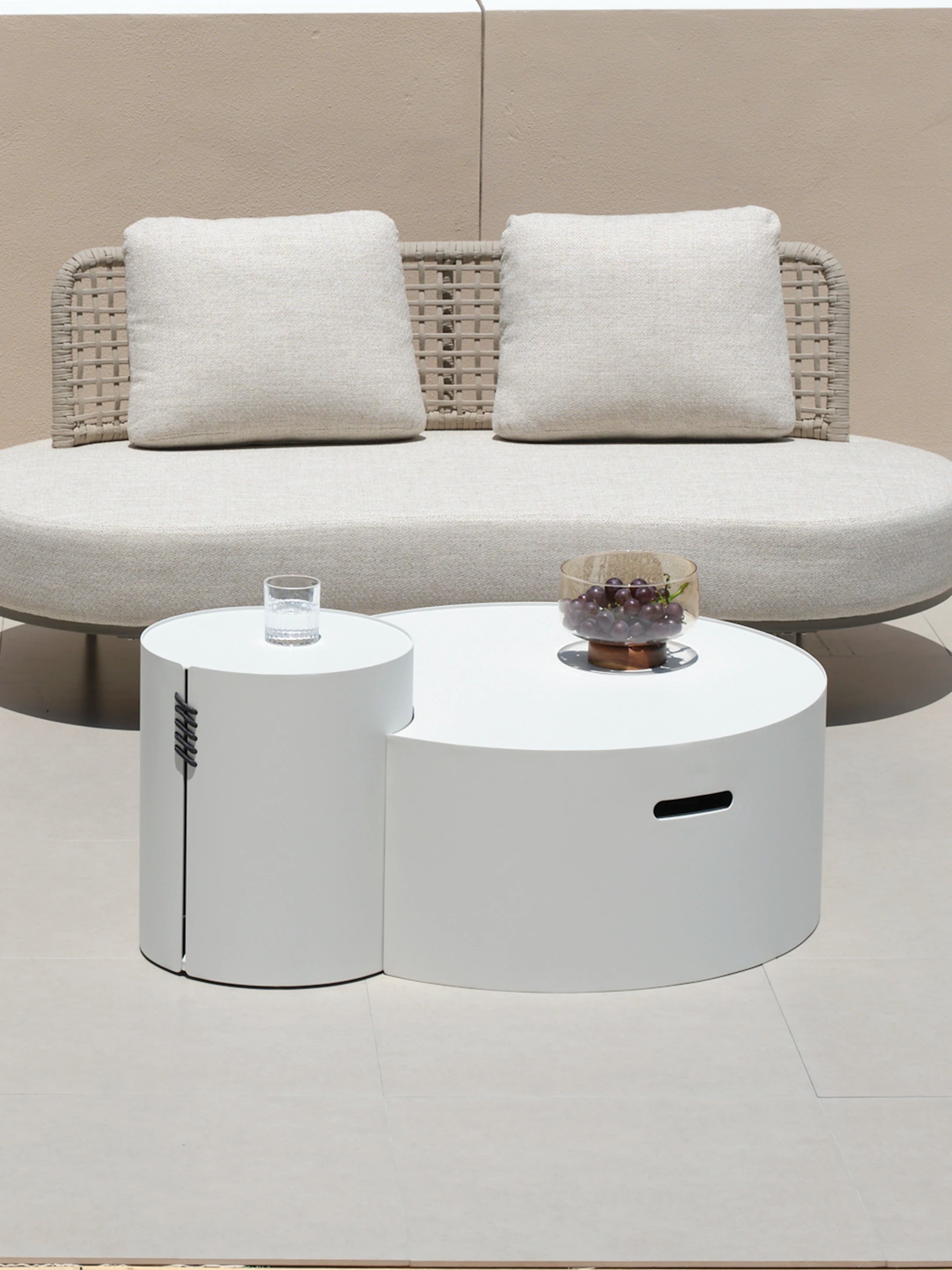 Speyer Crescent Couchtisch - Aluminium pulverbeschichtet-Weiß, bestehend aus zwei ineinandergreifenden runden Elementen in unterschiedlichen Höhen, ergänzt durch ein Glas Wasser und eine Schale mit dunklen Trauben darauf.