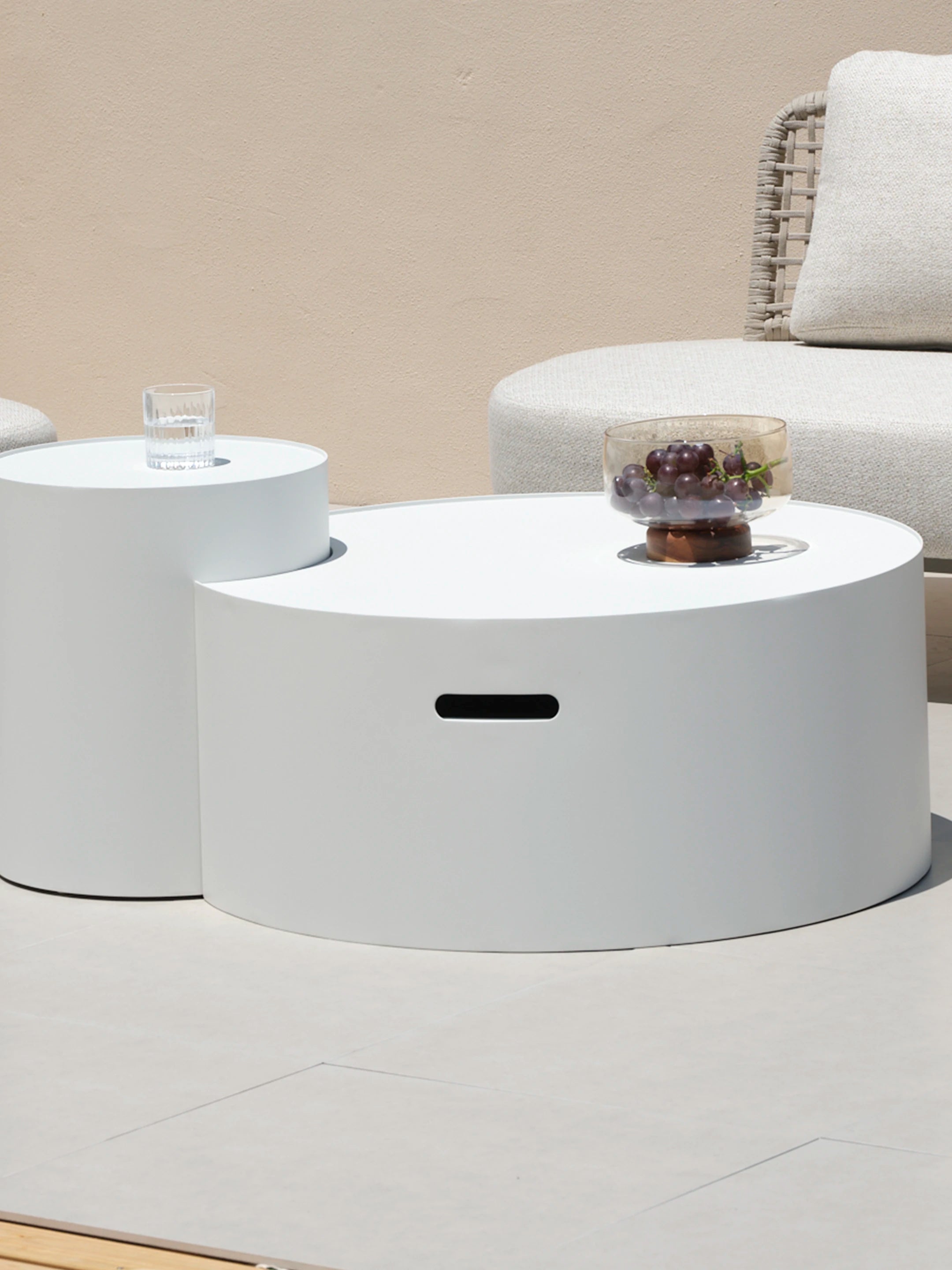 Speyer Crescent Couchtisch - Aluminium pulverbeschichtet-Weiß: Zwei unterschiedlich hohe, zylindrische Tische, einer mit Wasserglas, der andere mit Schale voller Trauben, vor einem gewebten Loungesessel und beigem Hintergrund.