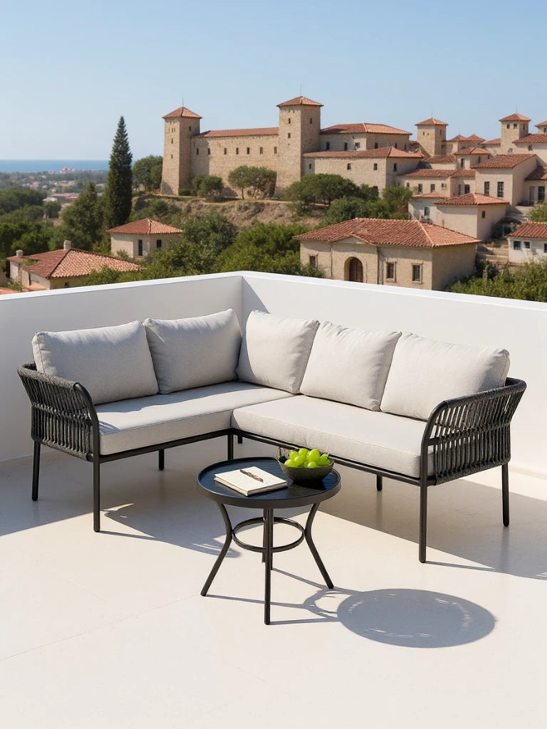 Malaga Garten Ecklounge Set (4-Sitzer) auf einer sonnigen Terrasse, mit lichtgrauen Kissen, rundem Beistelltisch mit Notizbuch und Äpfeln, im Hintergrund ein Dorf und das Meer sichtbar.
