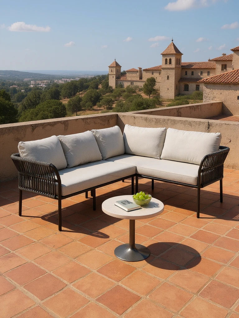 Malaga Garten Ecklounge Set (4-Sitzer) auf einer Terrasse: modernes L-förmiges Sofa mit dunkel gewebtem Rahmen, hellen Polstern, abgerundeten Armlehnen; kleiner Tisch mit Buch und Trauben.