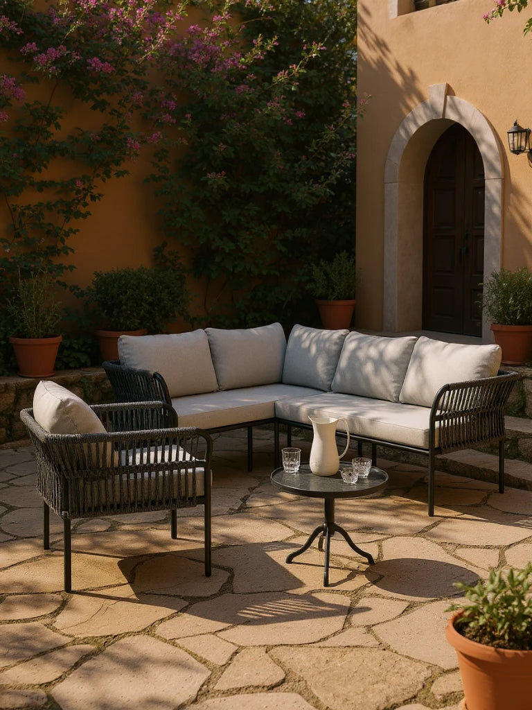 Malaga Garten Ecklounge Set (5-Sitzer) auf sonnenbeschienener Terrasse mit L-förmigem Sofa und Sessel, beide mit hellgrauen Kissen. Schwarzer Tisch mit weißem Krug, Gläsern, umgeben von Pflanzen.