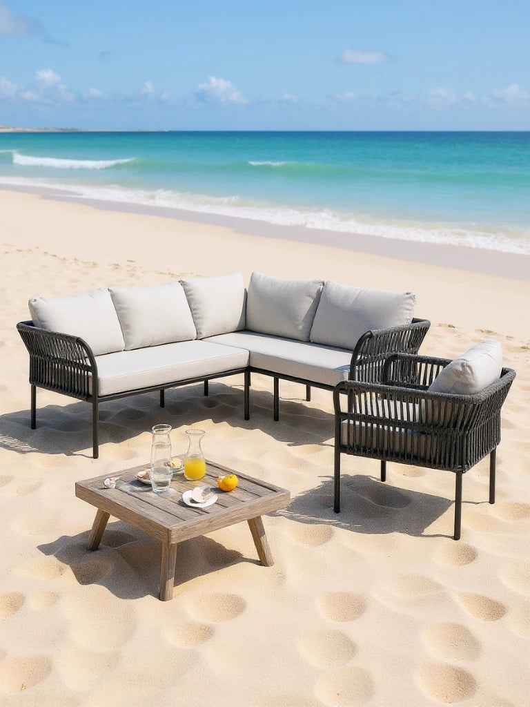 Malaga Garten Ecklounge Set (5-Sitzer) auf einer sandigen Terrasse: L-förmiges Sofa und Sessel mit schwarzem Rahmen, grauen Polstern, und quadratischer Holztisch mit Getränken und Muscheln.