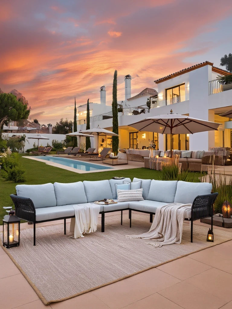 Malaga Garten Ecklounge Set (6-Sitzer) auf neutralem Teppich vor einer Villa bei Sonnenuntergang. L-förmiges Design mit blauen Kissen, handgefertigte Zugbänder, flankiert von Kerzenlaternen.