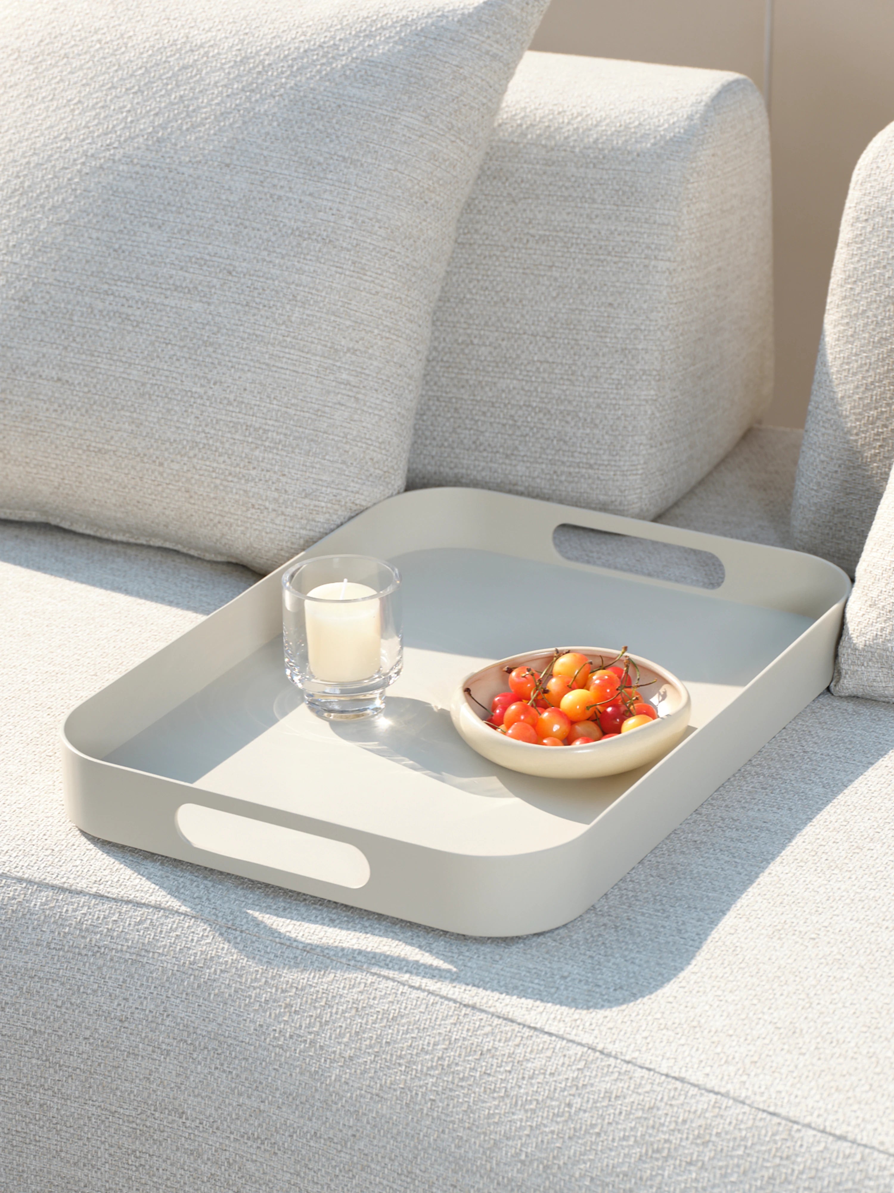 Palanga Tablett aus Aluminiumlegierung auf einem grauen Sofa, mit einem Glasvotiv und einer Schale Kirschen, beleuchtet von Sonnenlicht. Perfekt für stilvolle, wetterfeste Outdoor-Einrichtungen von Finqa GmbH.