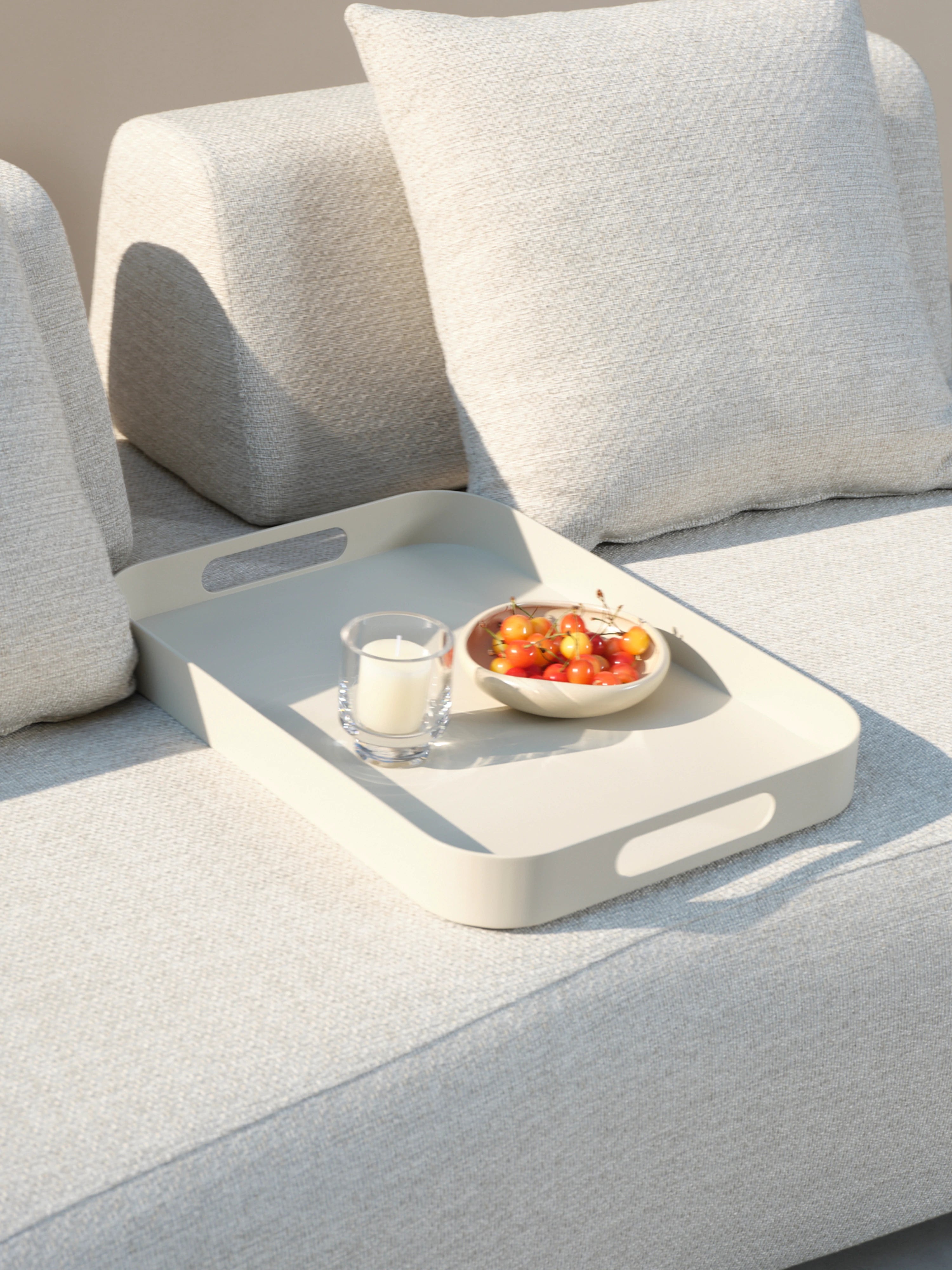 Palanga Tablett ruht auf einem hellgrauen Sofa, präsentiert mit einem Glasvotiv und einer Schale voller Kirschen. Perfekt für stilvolle Outdoor-Möbel von Finqa GmbH.