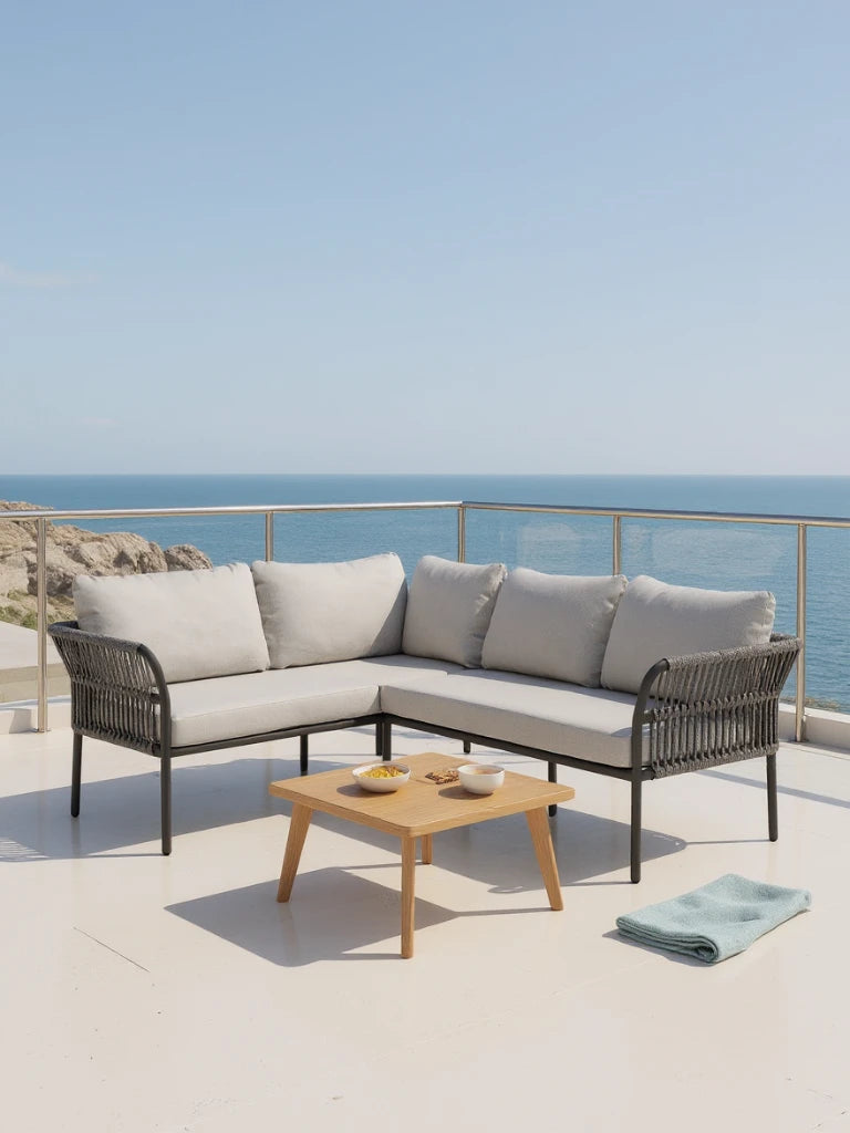 Malaga Garten Ecklounge Set (4-Sitzer) auf einer Terrasse, bestehend aus einem L-förmigen Sofa mit grauen Kissen und einem Holztisch, dahinter Meerblick. Ideal für stilvolle, wetterfeste Outdoor-Entspannung.