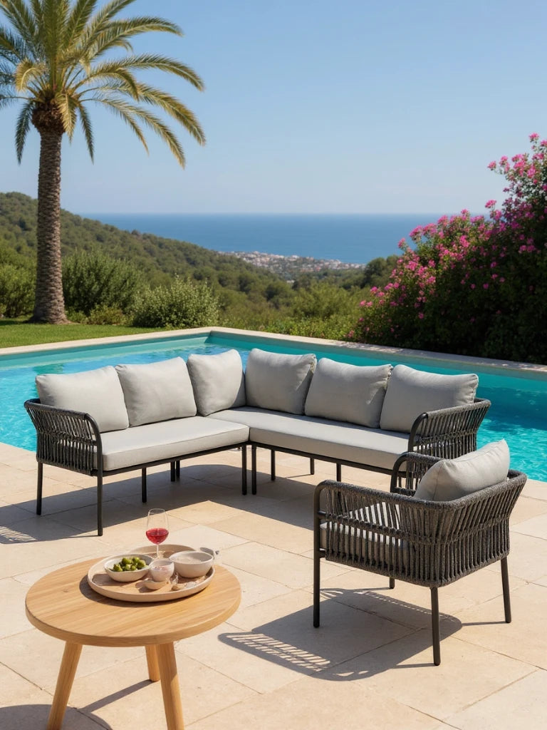 Malaga Garten Ecklounge Set (6-Sitzer) auf sonniger Terrasse mit grauen, gewebten Kissen, flankiert von einem passenden Sessel. Vorne steht ein runder Holztisch mit Snacks und Getränk.