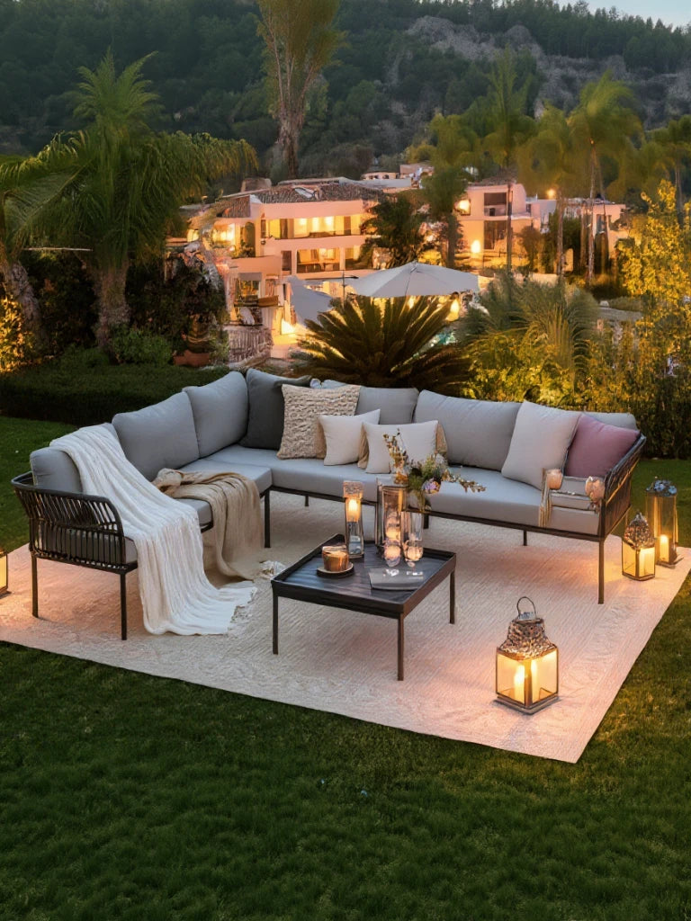 Malaga Garten Ecklounge Set (6-Sitzer) auf hellen Outdoor-Teppich: L-förmiges Sofa mit grauen Kissen, dekorativen Kissen und Decke, begleitet von einem niedrigen schwarzen Tisch mit Kerzenlaternen und Blumenarrangement.