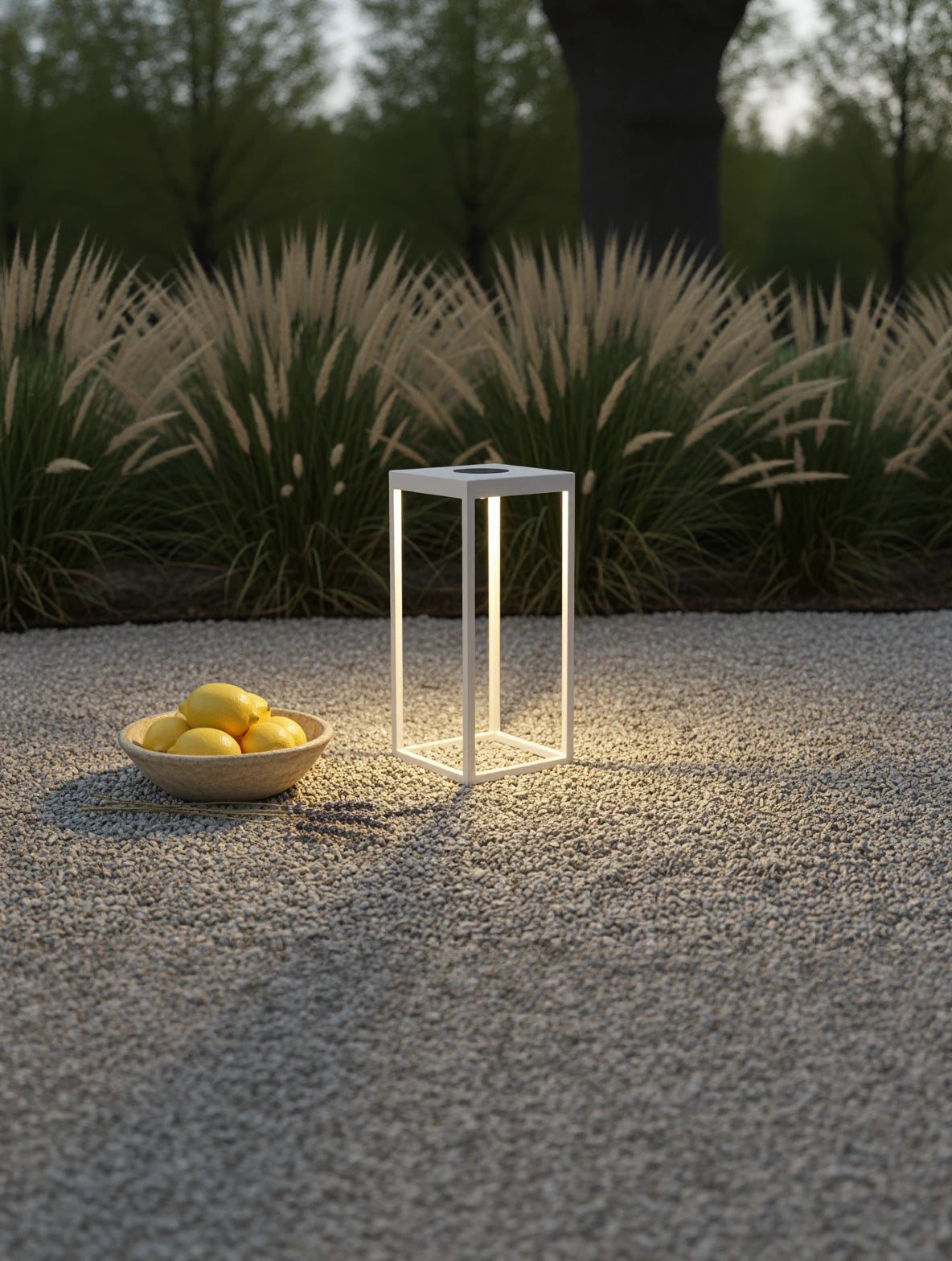 Kenya Solar Gartenlampe neben einer Schale mit Zitronen auf Kies, umgeben von Ziergräsern, strahlt warmes LED-Licht in einem minimalistischen, offenen Rahmen, ideal für abendliche Gartenatmosphäre.