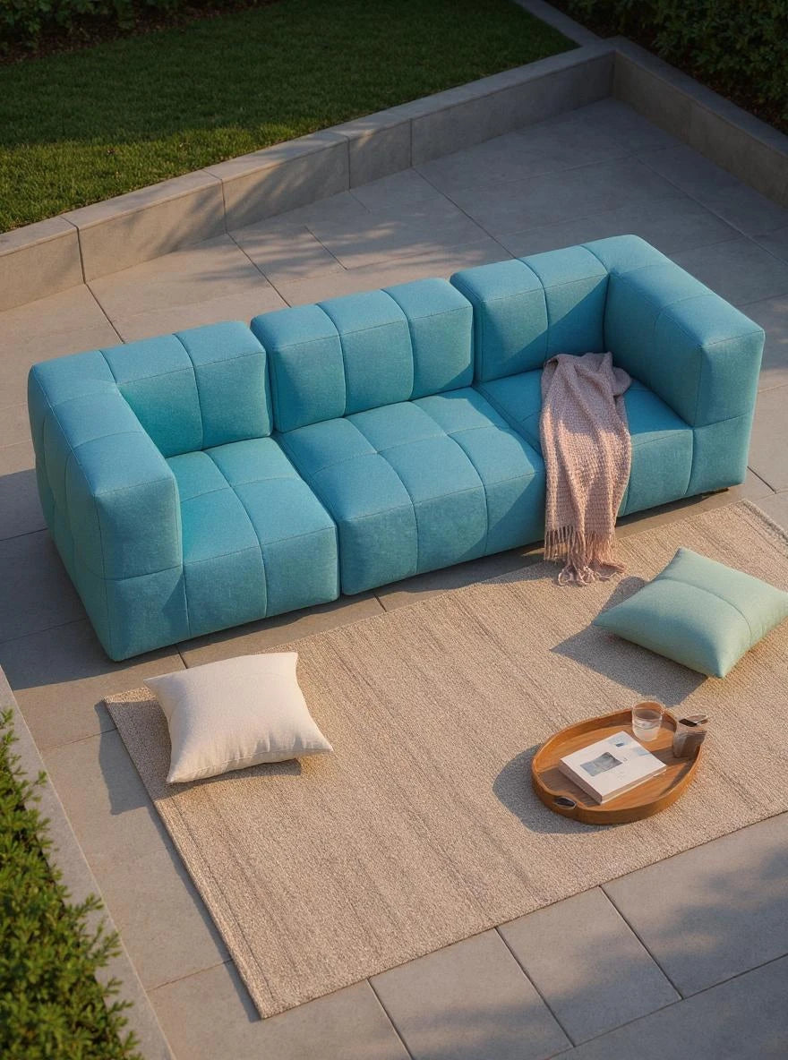 Modulares Garten-Lounge-Set Marshmallow, 3er-Set auf einer Terrasse: Teal-Sofa mit rosa Überwurf, zwei Bodenkissen, gewebter Teppich, Holztablett mit Glas und Buch, umgeben von gepflegter Bepflanzung.