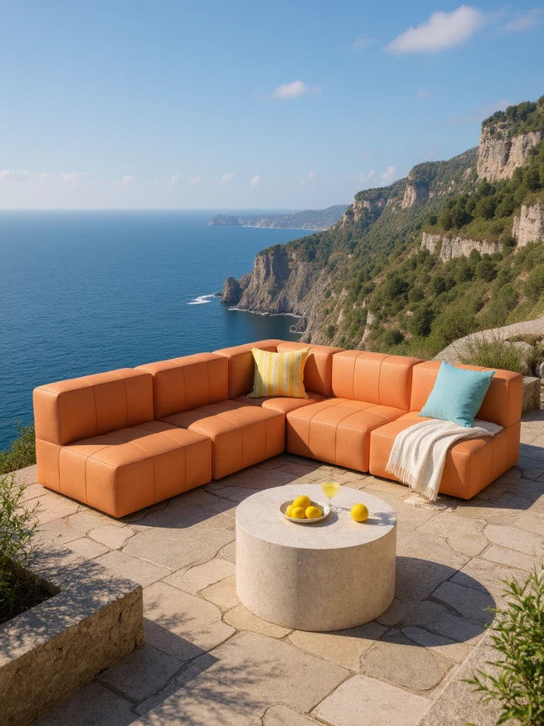 Modulares Garten-Lounge-Set Marshmallow, 5er-Set auf einer Küstenterrasse mit Aussicht. Orangefarbenes Sofa mit Kissen und Decke, begleitet von einem Steintisch mit Zitronen und Getränk.