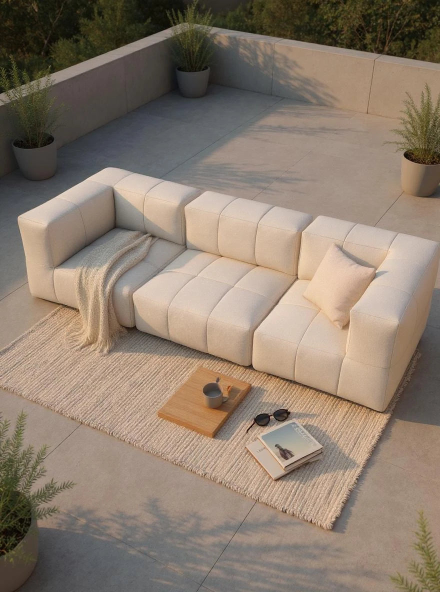 Modulares Garten-Lounge-Set Marshmallow, 3er-Set auf einer sonnigen Terrasse, bestehend aus einem cremefarbenen, modularen Sofa mit Kissen und Decke, Holzbrett mit Tasse, Magazin und Sonnenbrille auf dem Teppich.