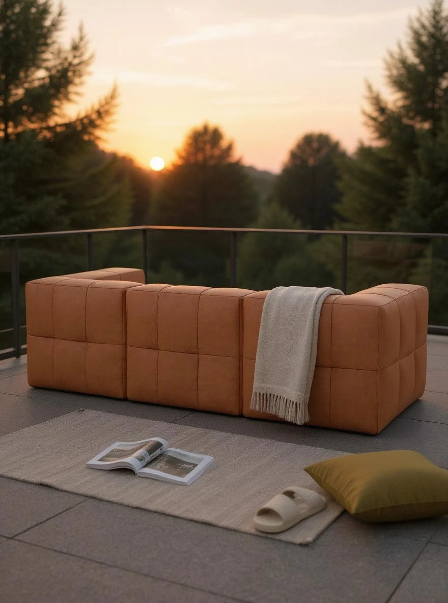 Modulares Garten-Lounge-Set Marshmallow, 3er-Set auf Terrasse bei Sonnenuntergang, bestehend aus drei kubischen Ledersofas, mit Decke, Buch, Pantoffel und Kissen auf Teppich davor.