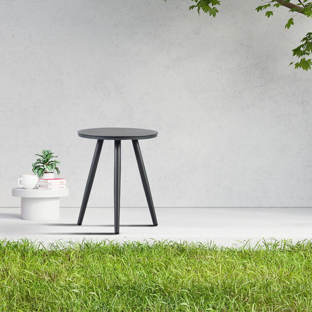 Finqa Prag Lounge Beistelltisch Aluminium-Pulverbeschichtung mit runder Tischplatte, elegant in einem minimalistischen Raum arrangiert, neben einem weißen Sockel mit Kaffeetasse, Büchern und einer Pflanze, unter Baumzweigen.