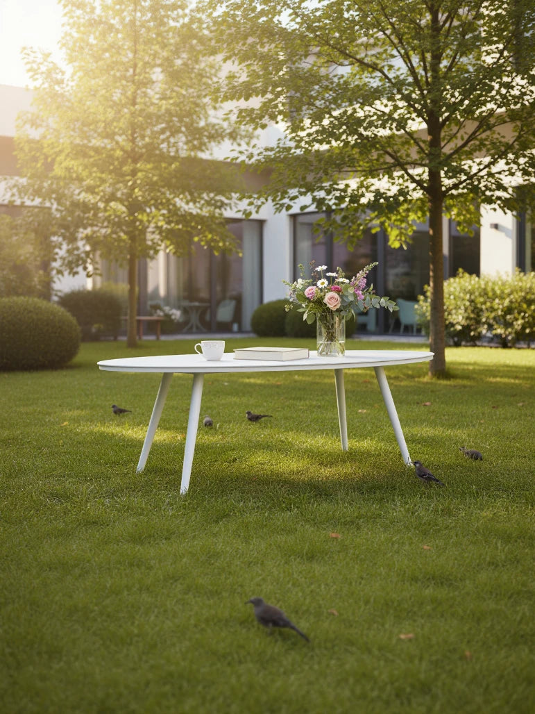 Freiburg Oval Boat Couchtisch aus pulverbeschichteter Aluminiumlegierung auf Rasen, mit Kaffeetasse, Buch und Blumenvase. Umgeben von Vögeln und Bäumen, ideal für stilvolle, wetterfeste Outdoor-Umgebungen.