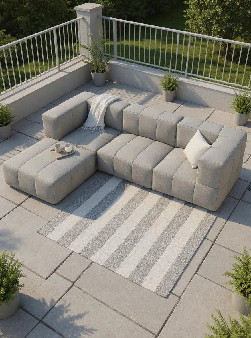 Das Bild zeigt das Babymax, 4er-Set, ein modulares, hellgraues L-förmiges Sofa mit Hocker auf einer Terrasse, ergänzt durch eine Decke, Kissen und stilvolle Pflanzenarrangements.