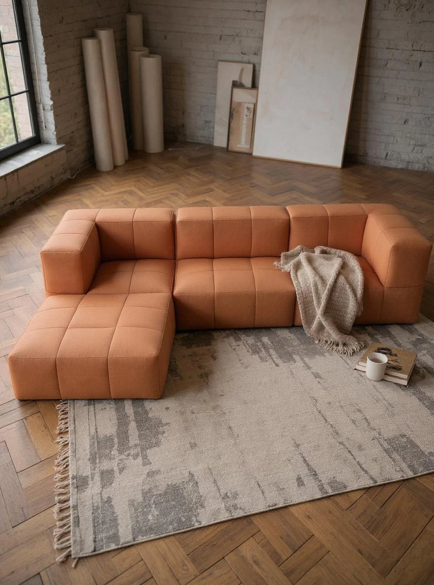 Babymax, 4er-Set: Modulares L-förmiges Sofa mit terrakottafarbener Polsterung, quadratisch abgesteppter Sitzfläche und Rückenlehne, im minimalistischen Loft-Stil, mit Bücherstapel und Tasse auf hell gemustertem Teppich.