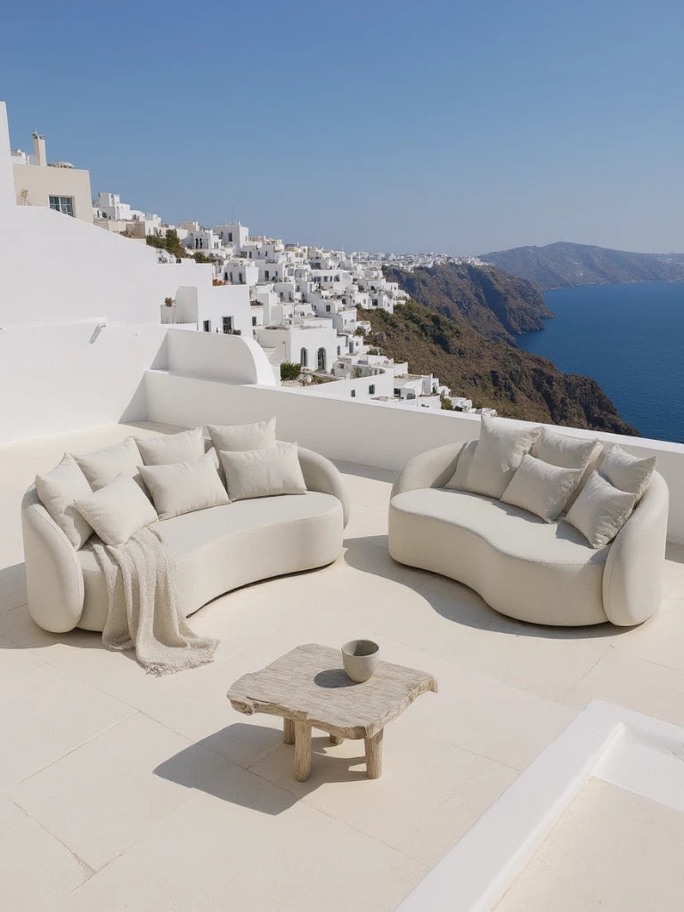 Santorini Garten Loungesofa Set (5-Sitzer) auf einer sonnenbeschienenen Terrasse, bestehend aus zwei geschwungenen, cremefarbenen Sofas mit Kissen und einem hellen Holztisch, überblickt das tiefblaue Meer.