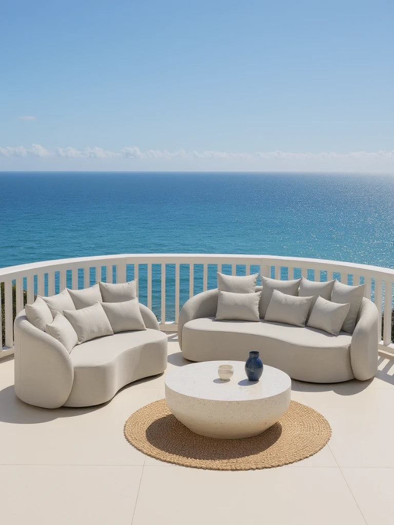Santorini Garten Loungesofa Set (5-Sitzer) auf einer Terrasse mit zwei geschwungenen, weißen Sofas und einem runden Couchtisch auf einem Jute-Teppich, mit Blick auf das Meer.